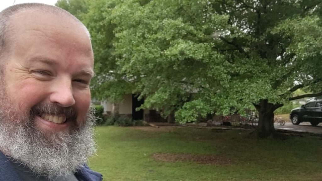 Fish Lee smiling, with a grey and white beard in front of a tree
