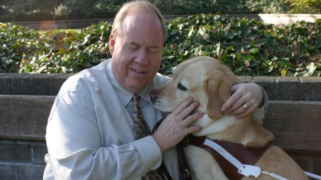 Michael Hingson wearing an off-white dress shirt with a gold and blue patterned necktie. Michael is shown feeling the face of his yellow Labrador retriever, Roselle.
