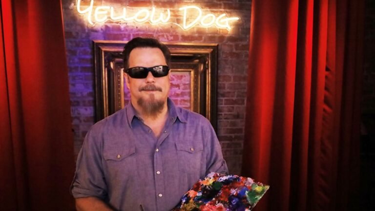A photo of John Bramblitt wearing black sunglasses and a blue shirt, holding an easel in front of a yellow sign reading "Yellow Dog". Photo Credit: Stephen Montoya: