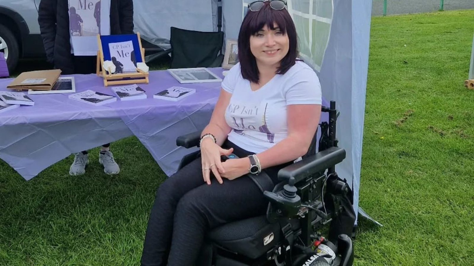 A picture of Samantha Maxwell in her power wheelchair smiling in front of a table.