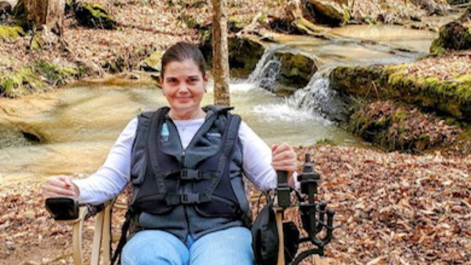 Melanie Dunn in her trackchair, wearing a green jacket and blue jeans, on a wooded trail.